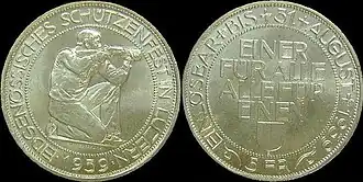 At left, the obverse of shooting thaler, showing a kneeling rifleman aiming to right, surrounded by the inscription EIDGENÖSSISCHES SCHÜTZENFEST IN LUZERN, with 1939 below. At right, the reverse, showing the words EINER FÜR ALLE, ALLE FÜR EINEN above the Lucerne coat of arms in the middle, with EINLÖSBAR BIS 31. AUGUST 1939 around the outer edge, and 5 FR at the bottom.