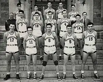 Three rows of five young men in white basketball uniforms stand on stone steps.