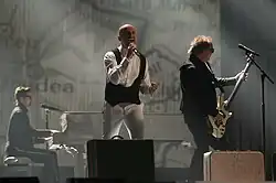 The picture sees a man singing into a microphone on a stage while wearing a black-white outift. He is accompanied by two fellow band members who play their instruments, and the LED in the background displays an oversized map of Romania.