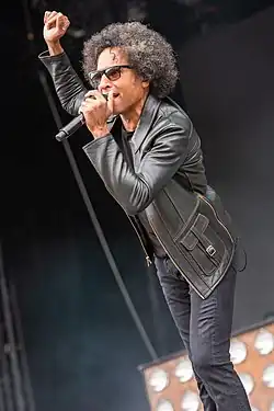 William DuVall wearing black jeans and a leather jacket, standing onstage, singing into microphone with one hand raised up