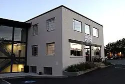 Photograph of a small, rectangular, two story brick building pained white. It is surrounded by an asphalt parking lot. It is annexed to a more modern glass and steel building with an open stairwell showing through the large windows. The sign on the front of the building reads, Kitchen and Bath Factory