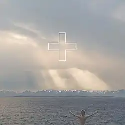 A man stands in front of a body of water and gazes into the sky with both of his hands up. A white outline of a plus sign is placed in the upper-center of the image