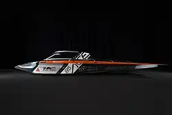 The image shows the streamlined solar race car called Silvretta, photographed from the side against a dark background. The car has a very low and flat profile, designed for maximum aerodynamic efficiency. Its body is primarily black and white, with orange accents, and is covered with various sponsor logos. The number “85” is clearly visible on the side. The cockpit is enclosed with a transparent canopy and is positioned toward the front-left side of the vehicle. On top is a layer of solar cells covering the whole car.