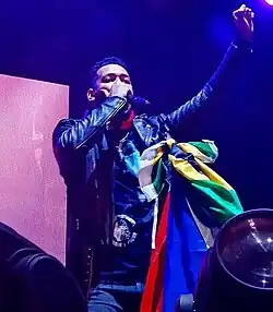 A man in a black leather jacket, with a microphone in his hand and the other raised high.