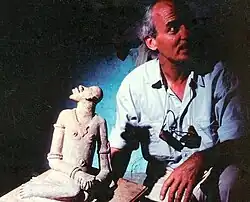 A balding white man, his face half obscured by shadow, poses next to a stone figurine.
