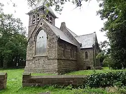 A photograph of a grey-bricked, many-windowed church with a slanted roof in a grassy field surrounded by green trees all under a bright sky