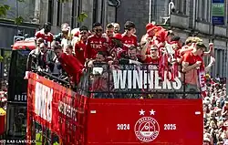 A football team in red outfits on top of an open air bus with the text Scottish Cup Winners 2024-2025 on the front of the bus. The bus is surrounded by cheering supporters.