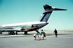 Rear three-quarter angle of twin-engined passenger jet with high tail plane, parked on tarmac with four people walking towards it