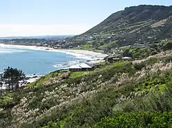 a sweeping beach with blue sea on the left and green hills on the right, small wooden dwellings dotted on the hills