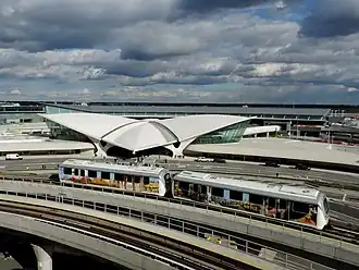An AirTrain vehicle in front of the TWA Flight Center. The vehicle is wrapped with advertisements and is traveling on a viaduct. Behind the AirTrain viaduct is the TWA Flight Center, a structure with a white roof shaped like a pair of wings.