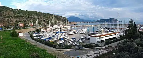 Dozens of sailboats crowd a marina under a cloudy sky along a mountain-lined coast.