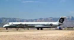 Right side view of an airplane taxiing on the ground towards left side of image. Another plane is behind it, and in the background are mountains and a blue sky with a few clouds.