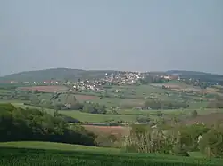 A small village with a visible church tower, on a low hill surrounded by fields and trees, seen from another such hill