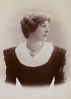 A white woman wearing a black dress with a white collar, and a chunky metallic necklace. Her shoulders are square to the camera but her face is almost in profile. Her dark hair is arranged back, away from her face and neck but covering her ears.
