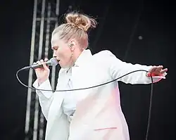 Ane Brun seen from left profile, wearing a white outfit, standing onstage with microphone held close to her mouth in right hand, appearing to sing, with eyes closed, and left hand holding microphone cord