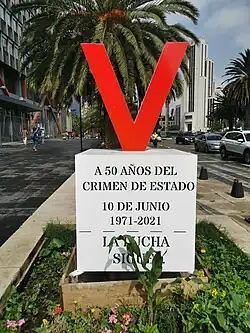A letter V is placed on top of a pedestal. The pedestal has written the phrases "50 years after the state crime", "10 June 1971–2021" and "The struggle continues!" in Spanish.