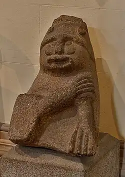 Photograph showing the torso and head of the Tandragee Idol, a grotesque and brutish figure with crossed arms in what appears to be a ritualistic pose. He has a gaping mouth, pierced nostrils and the stubs of what appear to be the ends of a horned helmet.