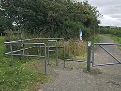 A metal barrier crosses the path, with a chicane and bar at ankle height.