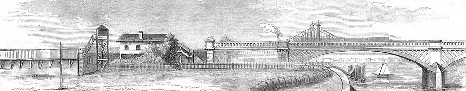 A railway line running (from left to right) on an embankment, past a wooden station building, over a girder bridge, and on to an arched girder bridge over a river