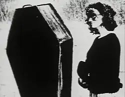 A masked woman standing beside an upright coffin