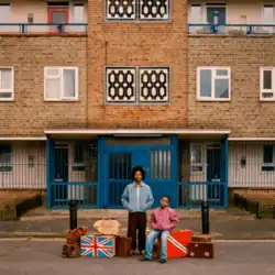Berwyn and a young boy surrounded by suitcases, two with the UK and Trinidadian flags on them, in front of a council estate building.