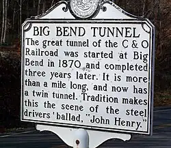 White plaque with the following text: BIG BEND TUNNEL. The great tunnel of the C & O Railroad was started at Big Bend in 1870 and completed three years later. It is more than a mile long, and now has a twin tunnel. Tradition makes this the scene of the steel drivers' ballad, "John Henry."