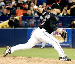 A man in a black baseball jersey, cap, and white pants pitches a baseball with his left hand.
