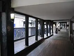 An elevated covered walkway with the fronts of premises on the right and posts and railings overlooking the street on the left