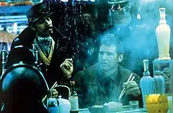 A man with close-cropped hair wearing a brown jacket sits at a counter, holding a pair of chopsticks poised over a rice bowl. Rain cascades down beside him as if from the edge of an awning. In the foreground is a teapot, several bottles, and other dining accessories. Steam or smoke rises from an unseen source. In the background, two standing men look down at the central figure. The goateed man on the left wears a dark snap-brim hat, a black coat with upturned collar, and a gold-trimmed vest. The man on the right, partly obscured by the steam, is wearing a constabulary-style uniform, featuring large wrap-around shades and a hat or helmet with a glossy, stiff brim. There is a bluish cast to the entire image.