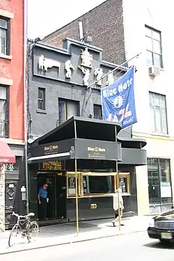 Street view in broad daylight of a two-story city building with a piano-shaped awning, gold-framed window panels at the bottom floor and jazz musician icons toward the roof, and a flag stemming from the top floor with the club's name; two pedestrians walk outside the building.