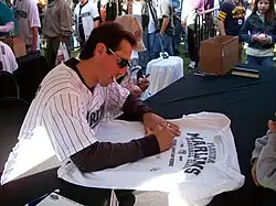 A young dark-haired man with sunglasses writing on a baseball jersey