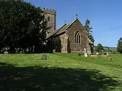 Stone building with square tower, partially obscured by trees. In the foreground are gravestones.
