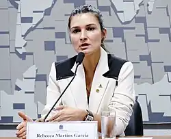A colour photograph of a woman speaking at a microphone, with a name label "Recbecca Martins Garcia" on the table before her