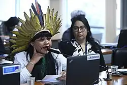 A woman is sitting behind a desk and speaking on a microphone. She is wearing a traditional head piece and there's another woman next to her.