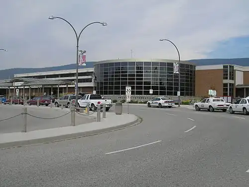 A road that contains vehicles parking along it, with a "Kelowna International Airport" sign in the background, as well as a building for an airport.