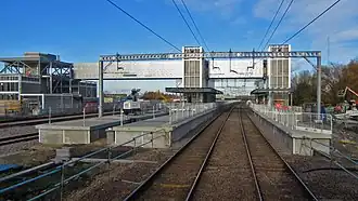 A picture taken at daytime from above the bed of two parallel tracks, in front of a single platform to the right and an island platform to the left. The platforms are built from concrete blocks and construction is still in progress. There is a footbridge at the back and a blue sky