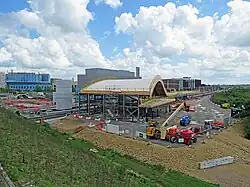 A panoramic view of a construction site, with the only clearly built structure a large concrete cuboid and an arched wooden roof structure suspended in the air by steel trusses. Also visible on the site are cherry pickers, scaffolding towers, other work vehicles and the two railway tracks running through the station.