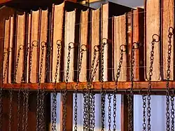 A bookshelf with 14 books. The page edges, not the spines, are facing the viewer. There are chains hanging from the middle of the edge of the covers.