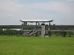 Exterior of Cheng Lim station, an elevated train station with a distinctive curved roof, surrounded by green grass and some distant trees under a partly cloudy sky
