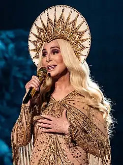 Cher sings on stage in a gold, beaded outfit with fringe detailing, wearing a large sunburst-style headpiece and long, platinum blonde hair.