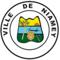 A coat of arms depicting a blue river between green land and a yellow sunset. A boat is on the river. A tree and a lion's head are on the land. The coat of arms is within two concentric white circles with black outlines, with the text "VILLE DE NIAMEY" in the outer circle.