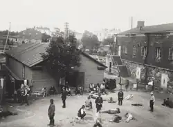 A vantage-point picture of a prison camp at the Suomenlinna Fortress in Helsinki. Around 25 Red prisoners are present in the courtyard, surrounded by a shack and a garrison building.