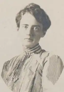 A white woman with dark hair in an updo, wearing a blouse with a high collar and frills