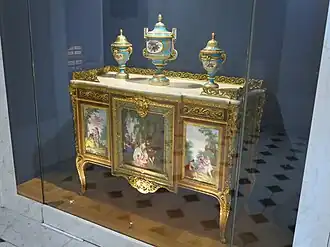 Louis XVI style commode of Madame du Barry; 1772; oak frame, veneer of pearwood, rosewood and kingwood, soft-paste Sèvres porcelain, gilded bronze, white marble, and glass; height: 0.87 m, width: 1.19 m, depth: 0.48 m[105]