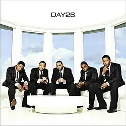 The cover shows the group dressed in black suits with neckties, sitting on a white couch near a table. Behind the group is a blue sky with clouds. The band's name is featured on the ceiling, colored in black.