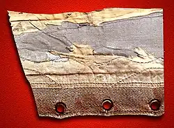 A piece of badly distressed fabric on a red background. The fabric is crossed by three seams and there are three eyelets at the bottom of it.