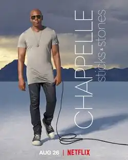 A bald man wearing sunglasses and a long grey tshirt and black pants, stands holding a microphone. He is outside on the plain white ground of the Bonneville Salt Flats and mountains can be seen in the distance.