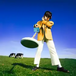 McKenna in a yellow coat standing in a green paddock holding a metal detector to the ground, with two cows in the background.