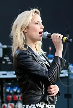 A blonde woman in a leather jacket and floral dress holding a microphone.