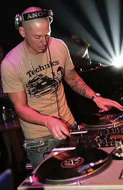 DJ Rectangle wearing headphones and a Technics t-shirt while mixing on turntables during a live DJ set, with stage lights and equipment visible.